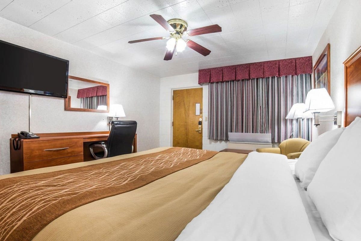 Habitación de hotel con cama, escritorio con teléfono, silla, ventilador de techo, televisor montado en la pared y ventana con cortinas.