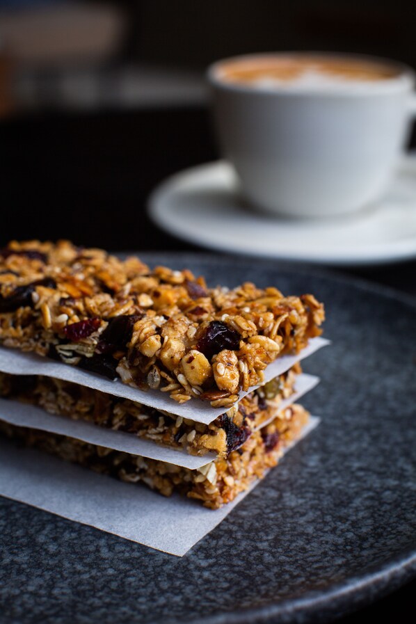 Unas barras de granola apiladas sobre un plato oscuro, con una taza de café al fondo.