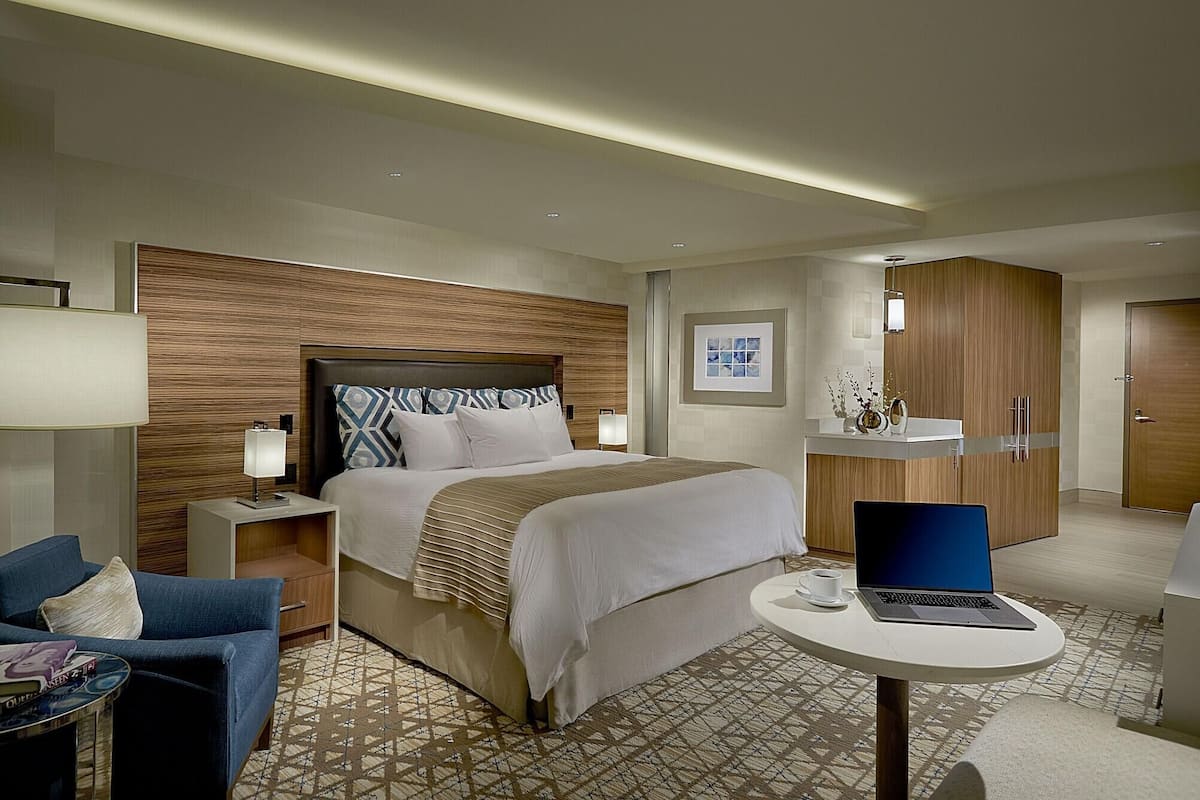 A hotel room with a bed, a desk with a laptop, a chair, a bedside table with a lamp, and a wardrobe.