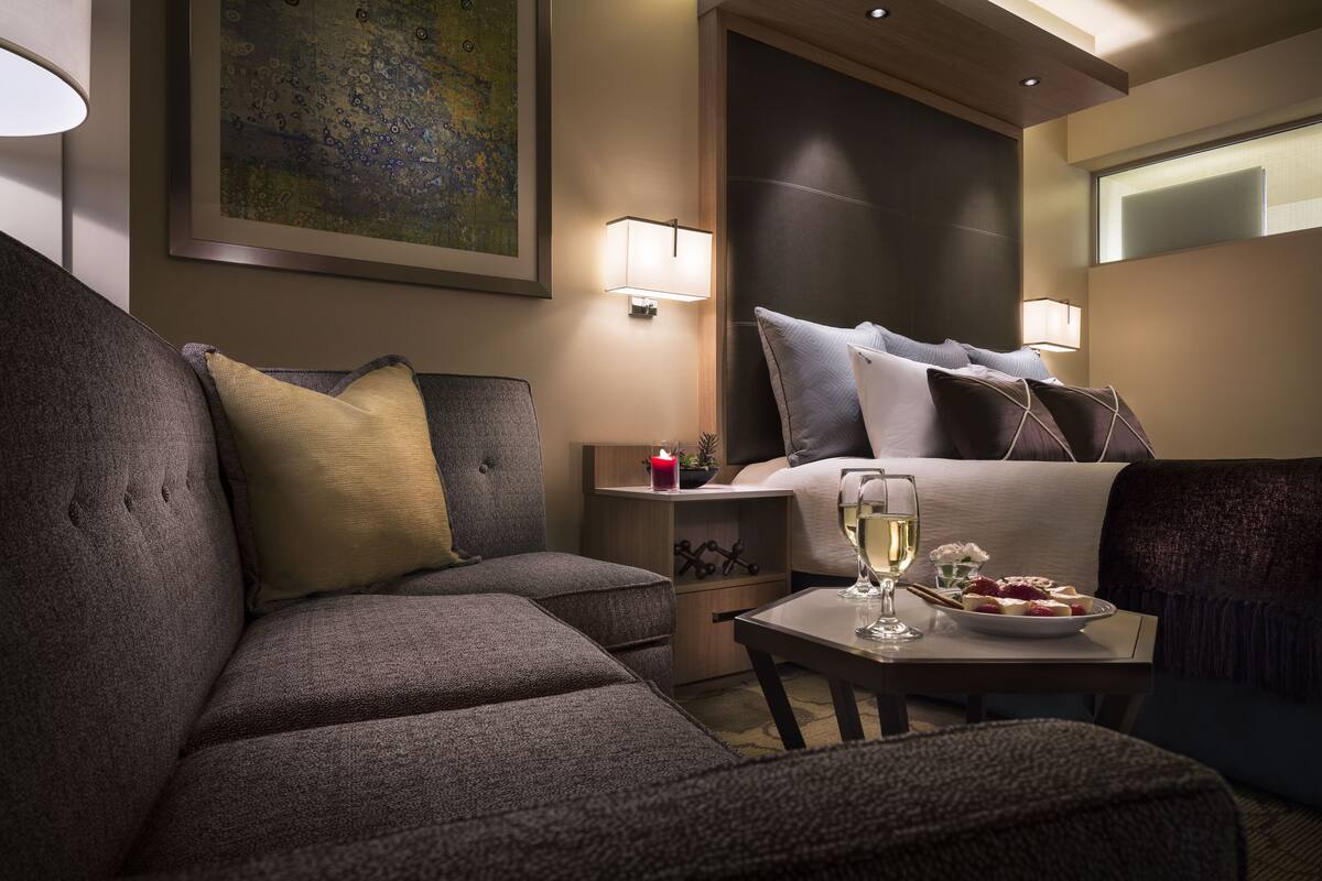 Une chambre d’hôtel avec un grand lit, un canapé, une petite table avec un verre de vin et une assiette de fruits, et une table de chevet avec une lampe.