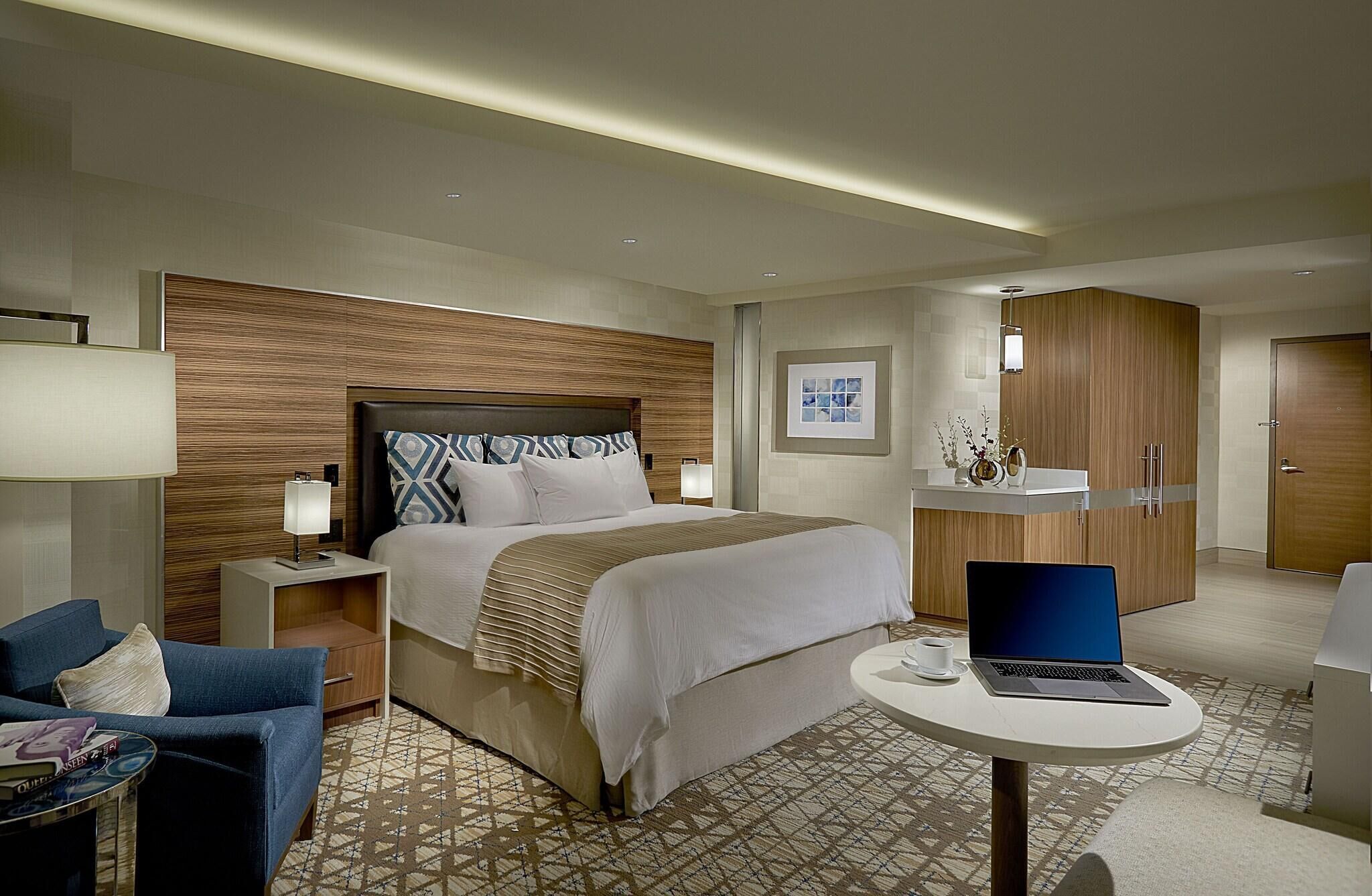 Una habitación de hotel con cama, escritorio con computadora portátil, silla, mesita de noche y lámpara.