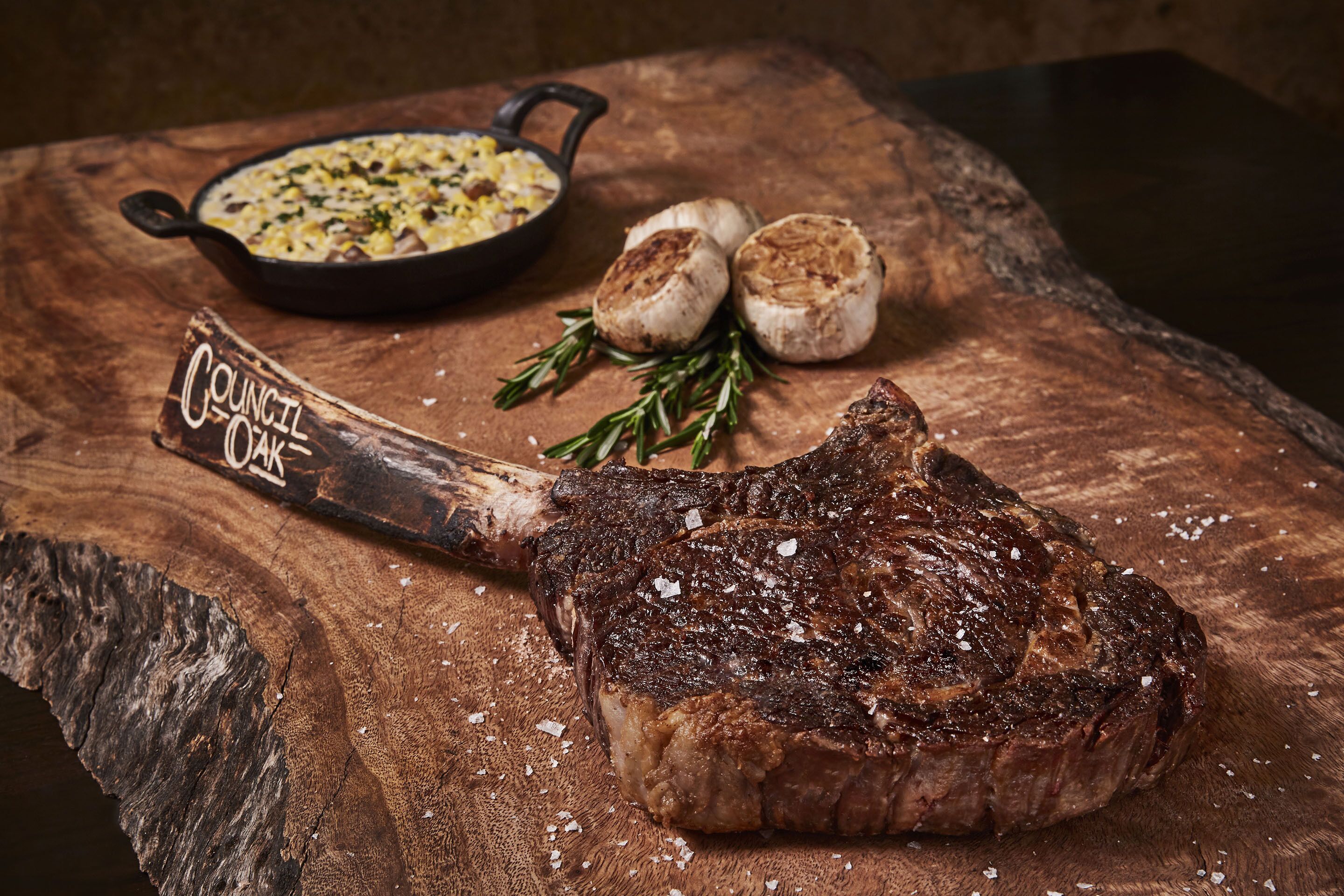 Un steak avec os, servi avec une poêlée d’accompagnements et deux pommes de terre rôties sur une surface en bois.