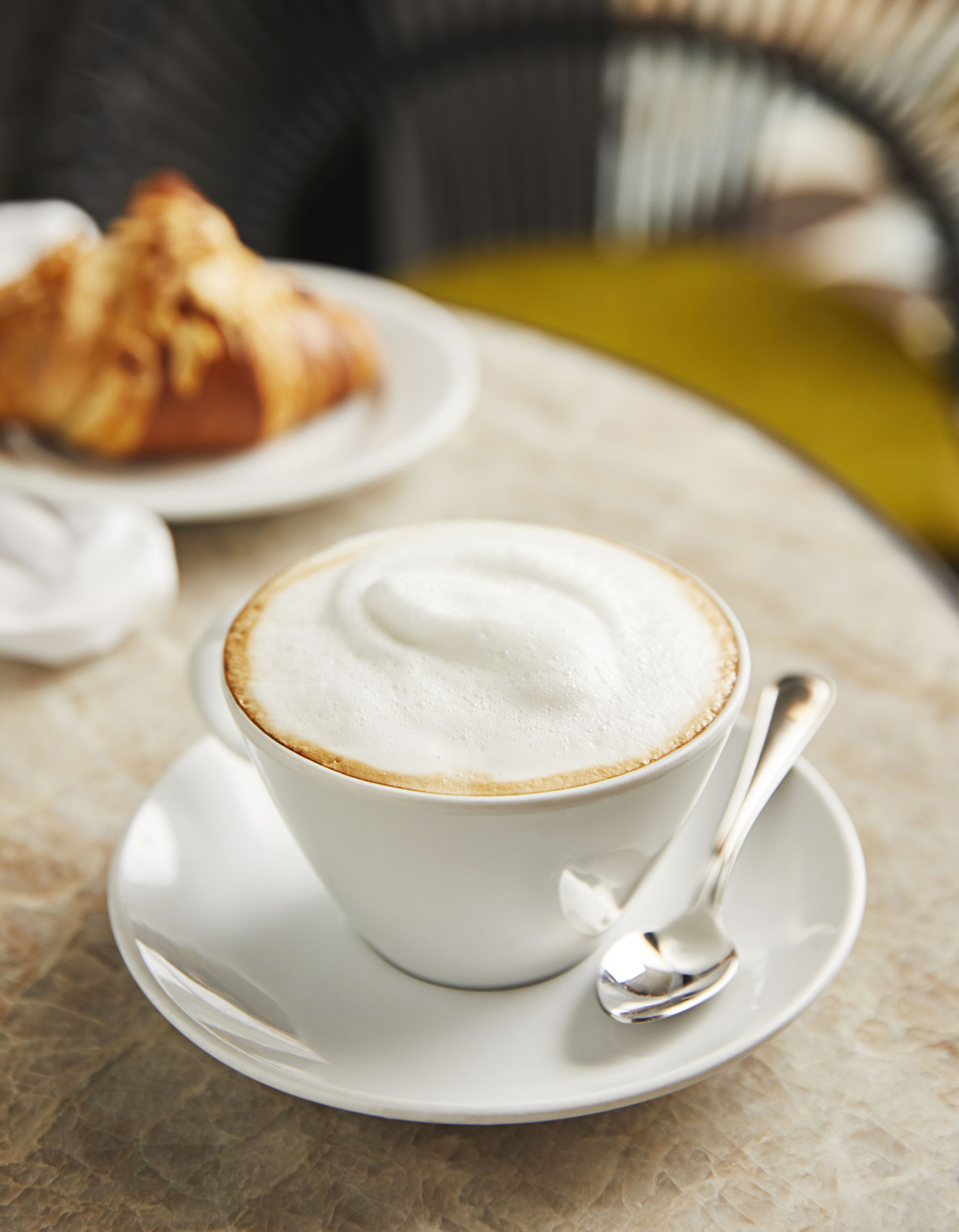 Een kop koffie met schuim op een witte schotel, met een lepel, op een marmeren tafel.
