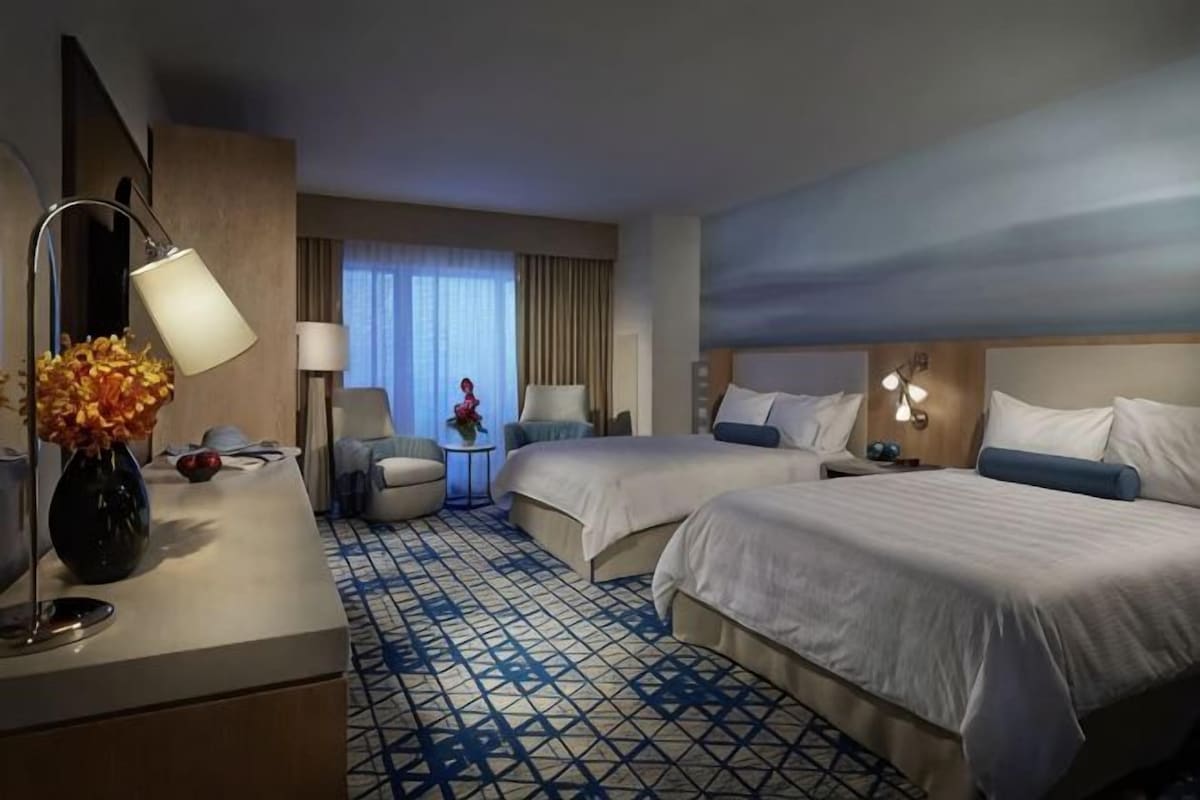 A hotel room with two beds, a desk, a chair, a lamp, and a vase with flowers.