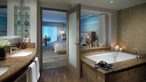 A modern hotel bathroom with a large sink, a bathtub, and a separate shower area. There is a bed visible in the adjacent room.