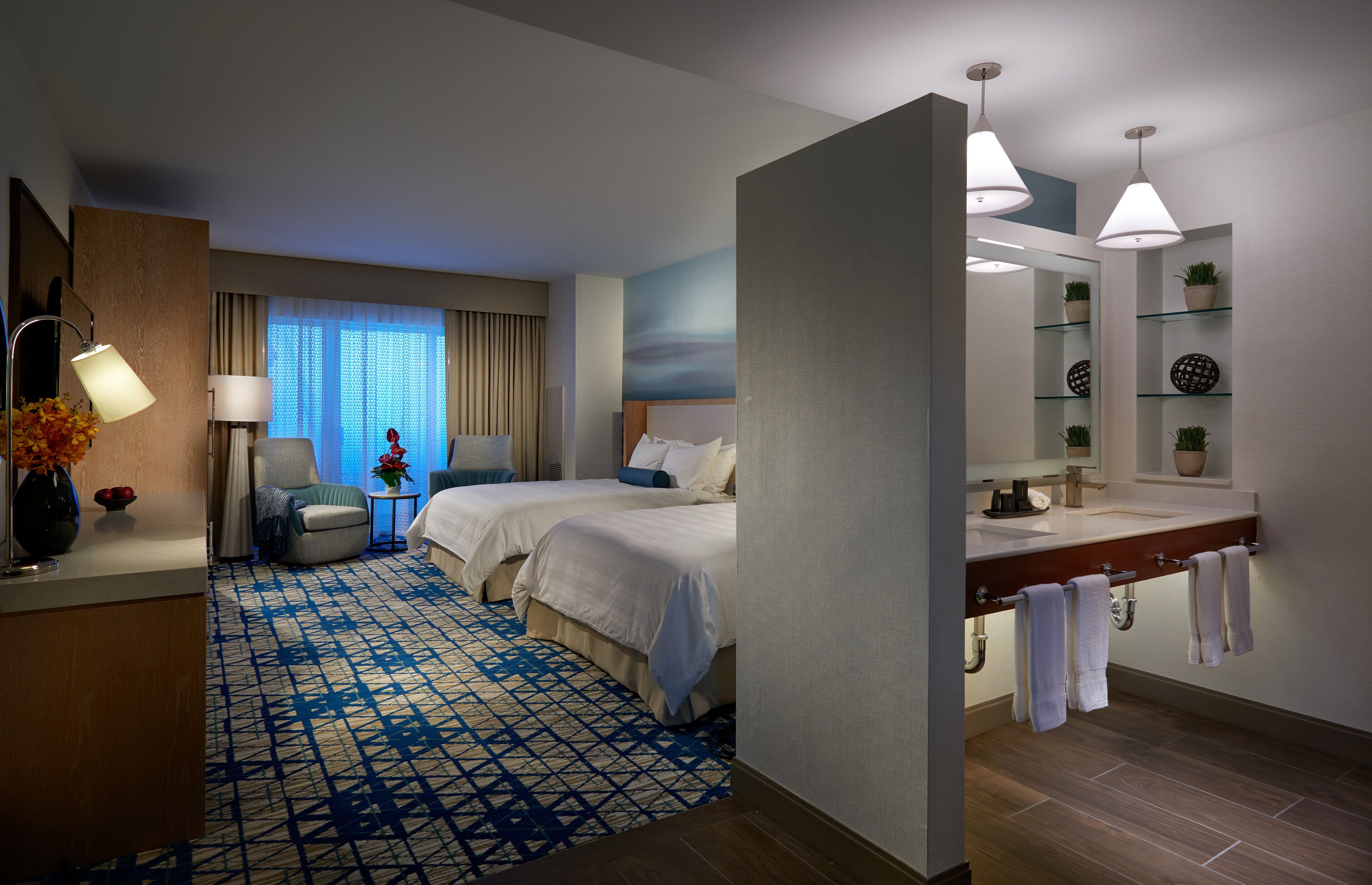 Habitación de hotel con dos camas, una zona de estar con una silla, un escritorio, un amplio ventanal con cortinas, un baño con lavabo y espejo, y una alfombra estampada.