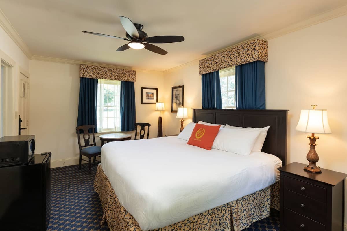 A hotel room with a bed, a desk, a chair, a TV, a lamp, and a ceiling fan.