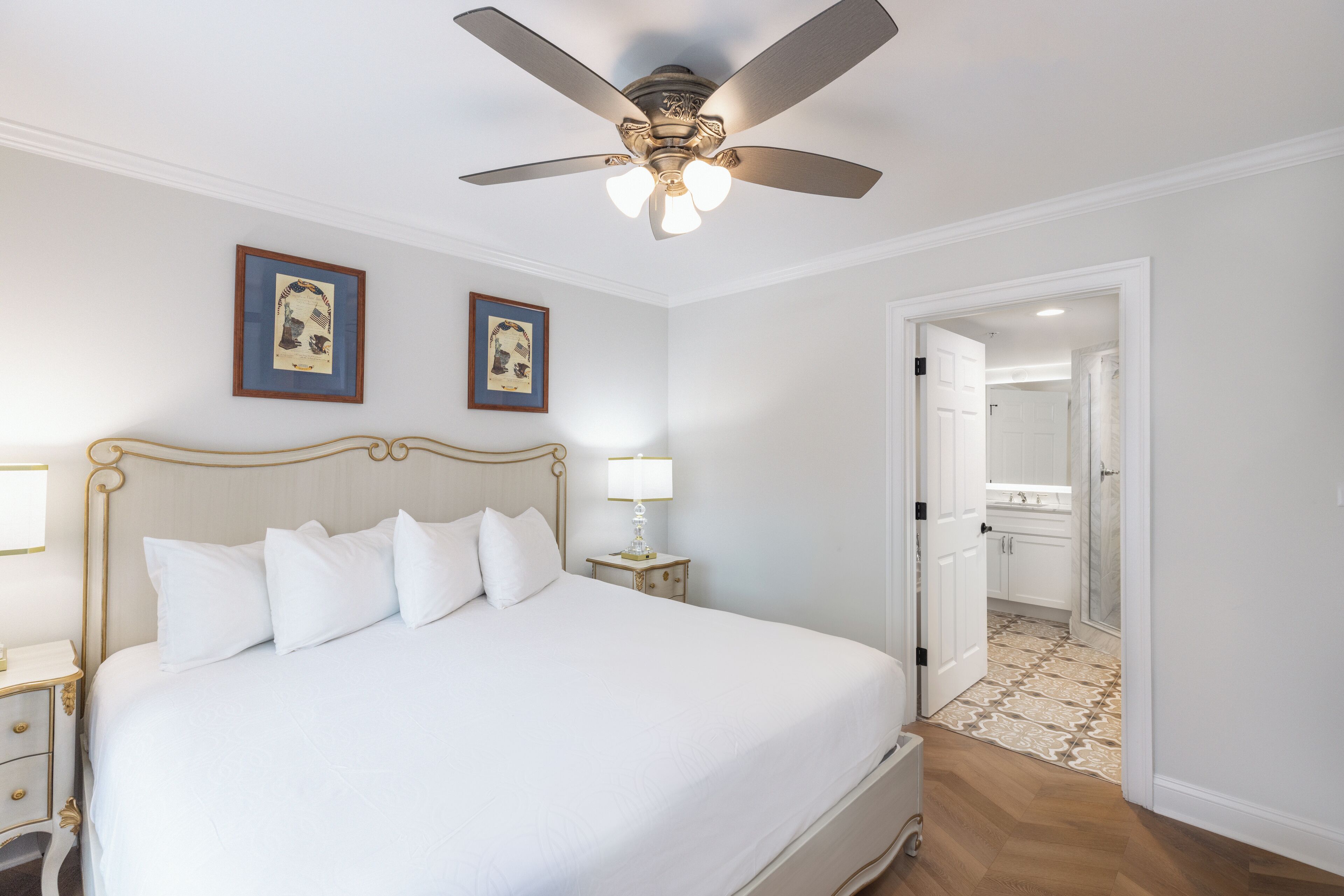 Un dormitorio ordenado con una cama grande, un ventilador de techo y cuadros en la pared.