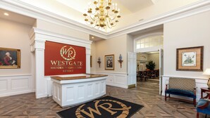 Een hotel lobby met een receptiebalie, een kroonluchter, ingelijste kunstwerken en een vloer met een patroon.