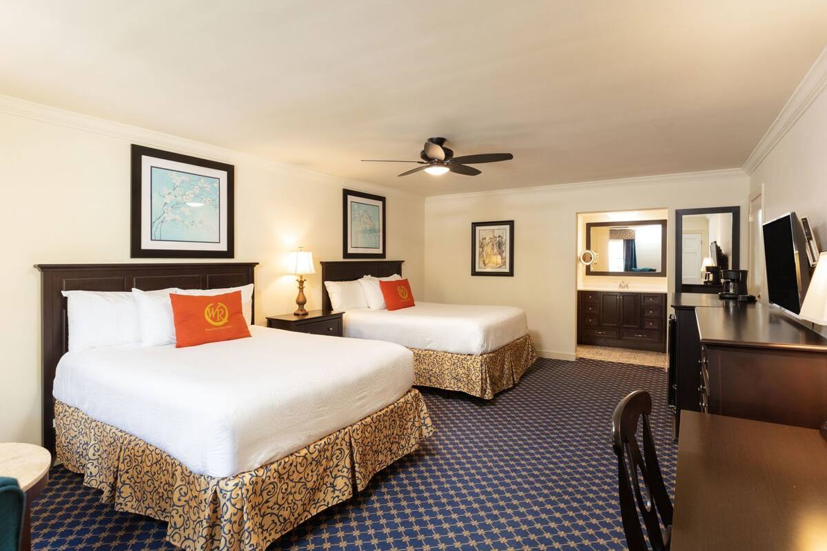 A hotel room with two beds, a desk, a chair, a ceiling fan, and a bathroom with a mirror and sink.