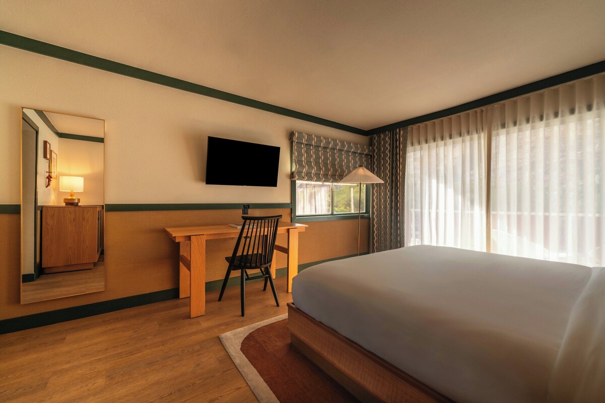 Habitación de hotel con cama, escritorio, silla, televisor y ventana con cortinas.