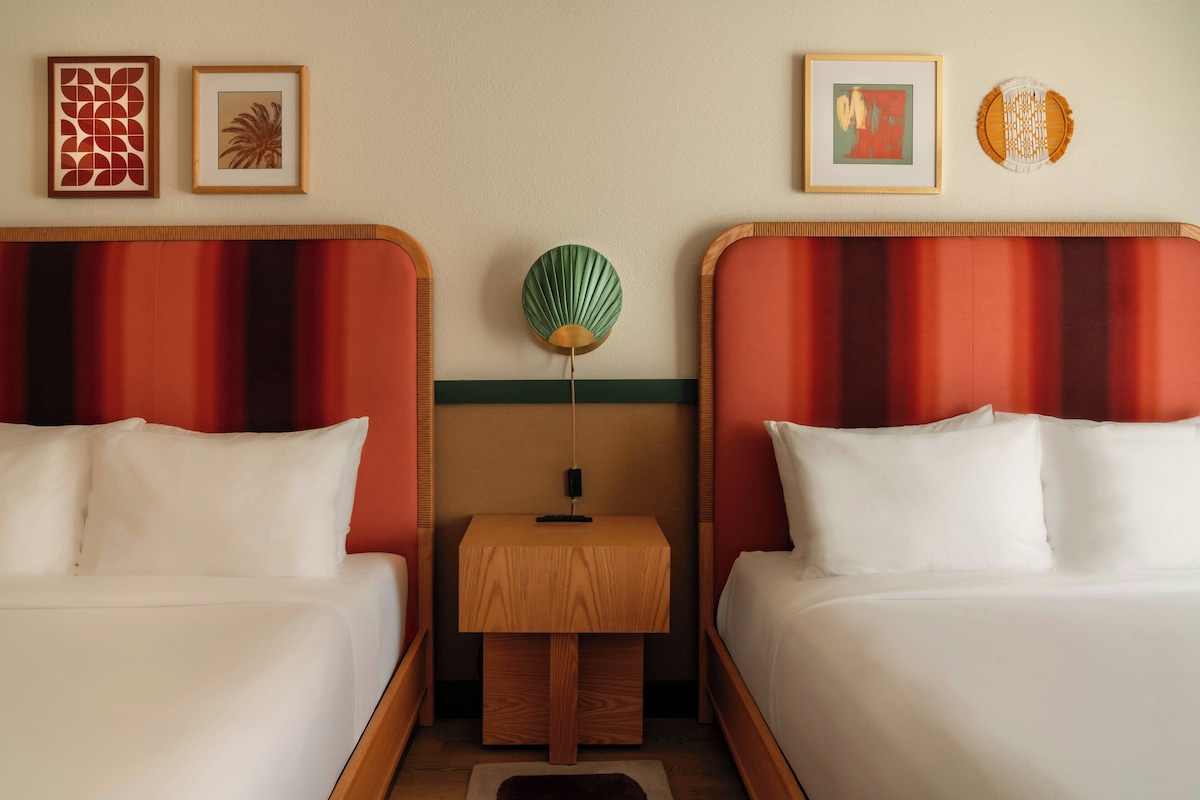 A hotel room with two beds, a nightstand, and a wall-mounted lamp.