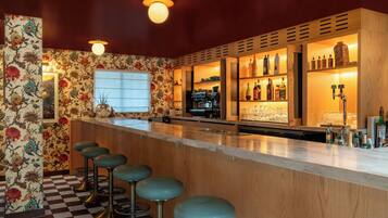 Un área de bar con suelo a cuadros, mostrador de madera, taburetes y una variedad de botellas y vasos.