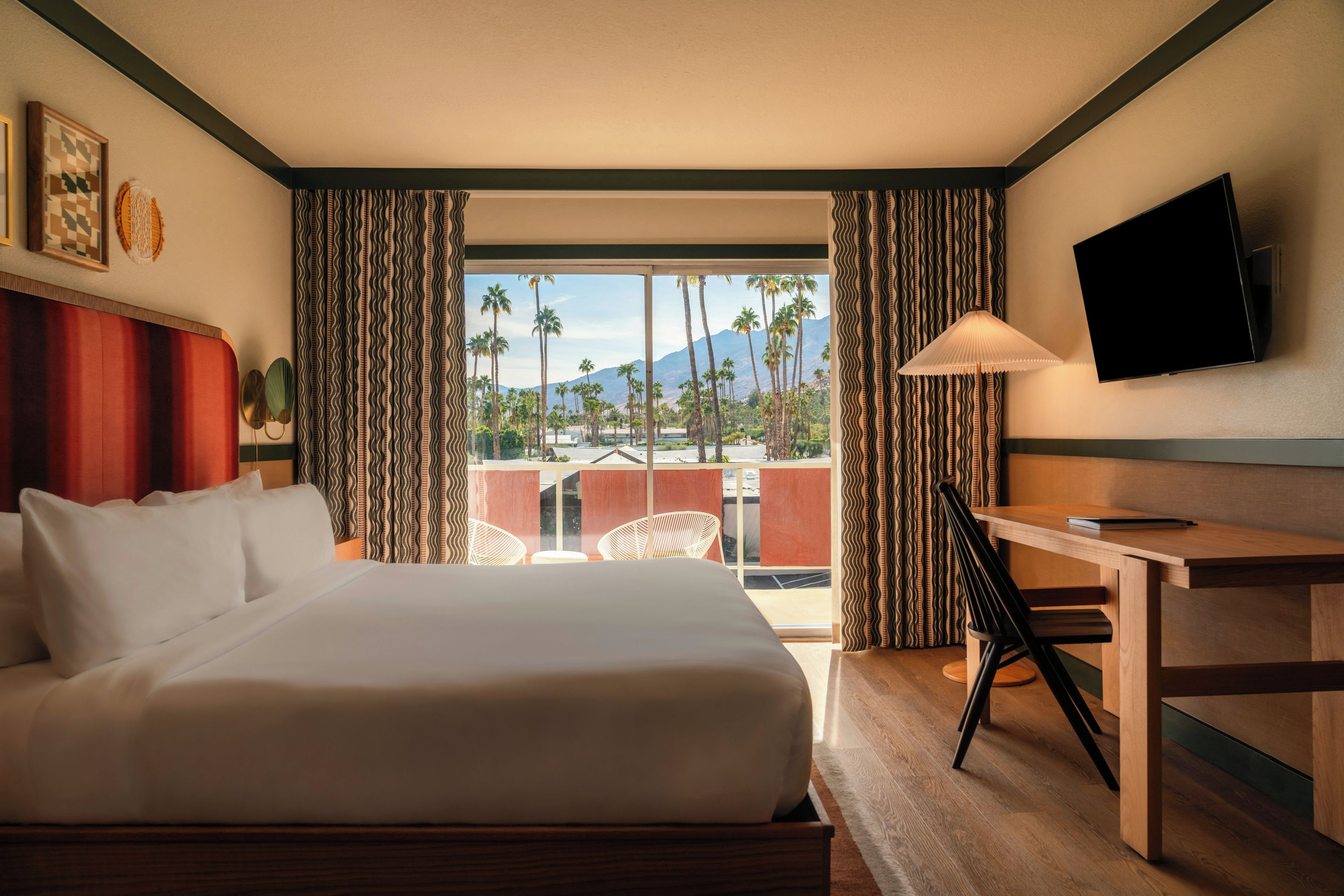 A hotel room with a large bed, a desk, a television, and a view of palm trees and mountains.