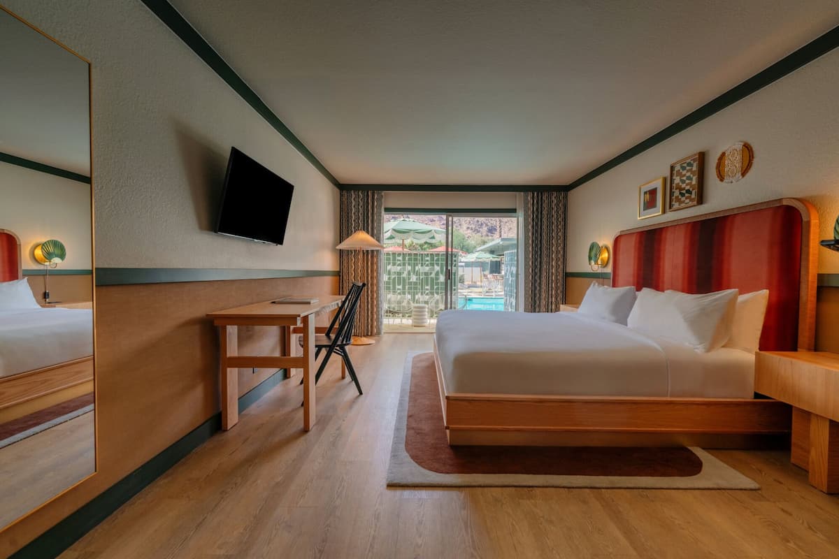 A hotel room with a bed, a desk, a chair, a TV, and a view of an outdoor pool area.