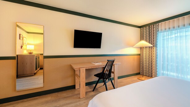 A hotel room with a bed, a desk, a chair, a TV, and a window with curtains.