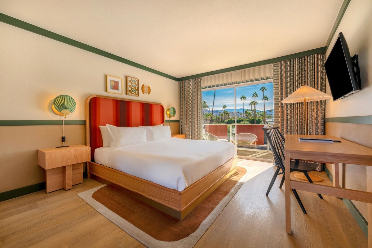 A hotel room with a bed, a desk, a chair, a TV, and a view of palm trees.