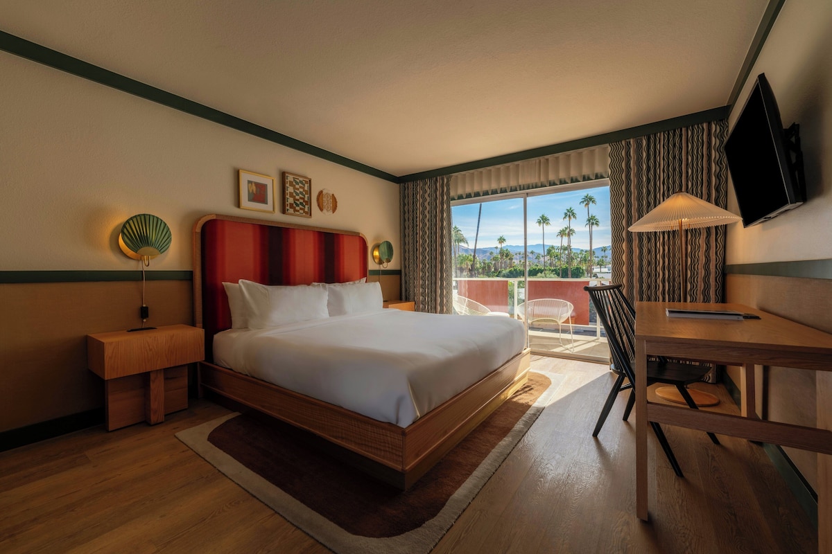 A hotel room with a bed, a desk, a chair, a TV, and a balcony with a view of palm trees.
