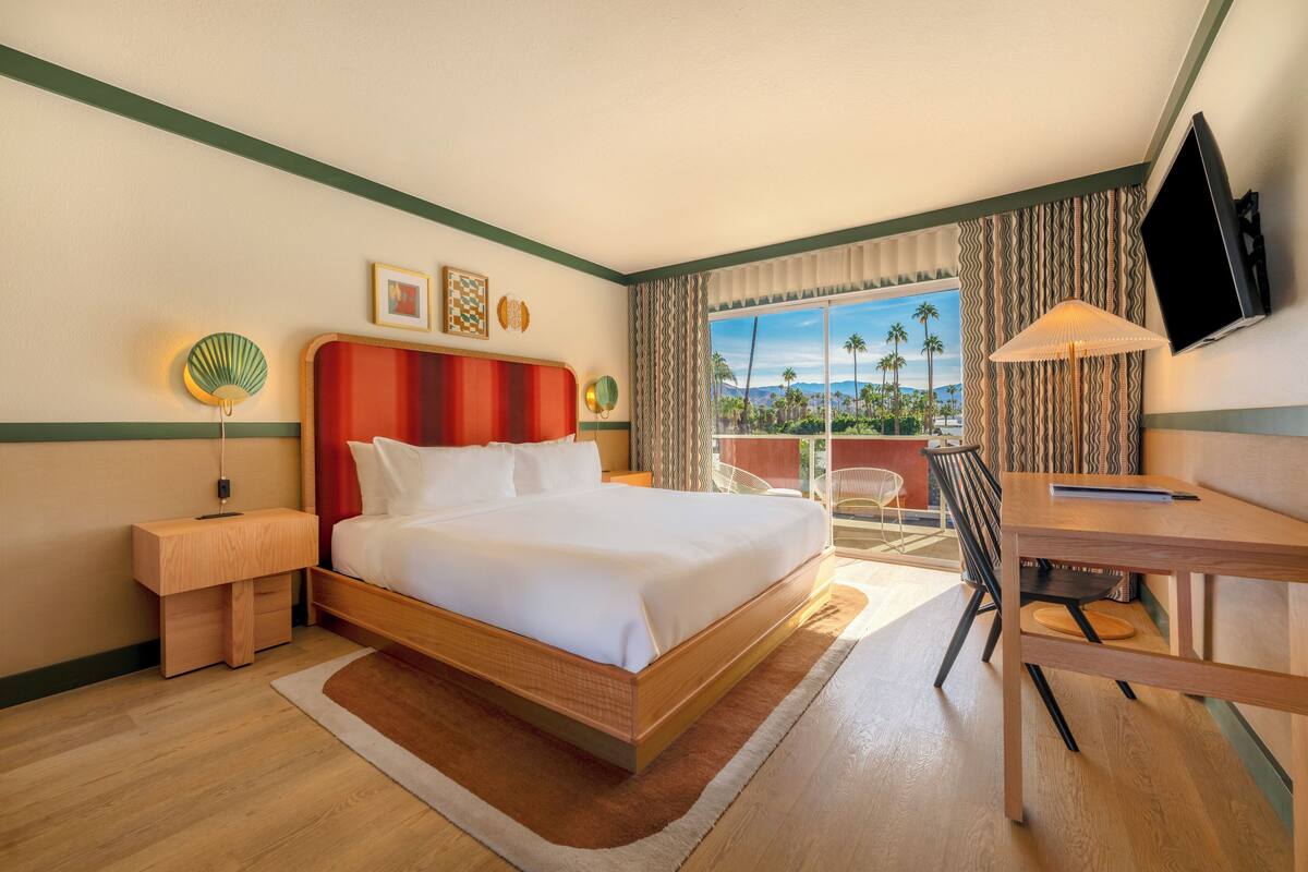 A hotel room with a bed, desk, and a view of palm trees.