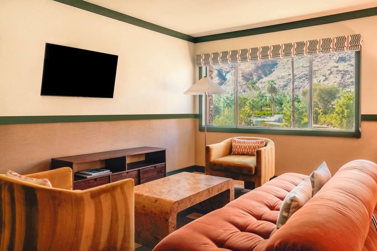 A living room with a flat-screen TV mounted on the wall, a sofa, a coffee table, and a window with a view of greenery and mountains.