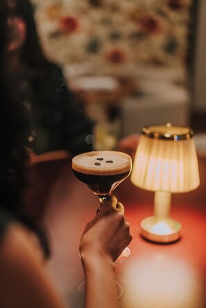 A hand holding a martini glass with a frothy top, a lit table lamp, and a blurred background with a floral pattern.