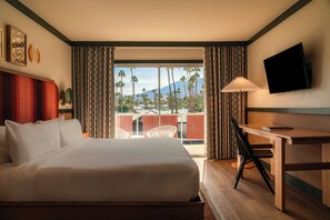 A hotel room with a large bed, a desk, a television, and a view of palm trees and mountains.