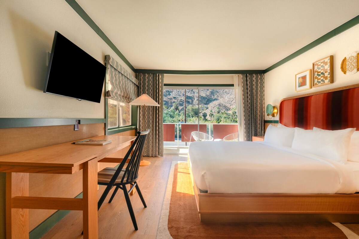 A hotel room with a bed, desk, chair, TV, and a balcony with a view.