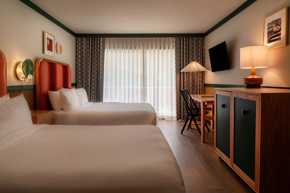 A hotel room with two beds, a desk, a chair, a television, and a lamp.