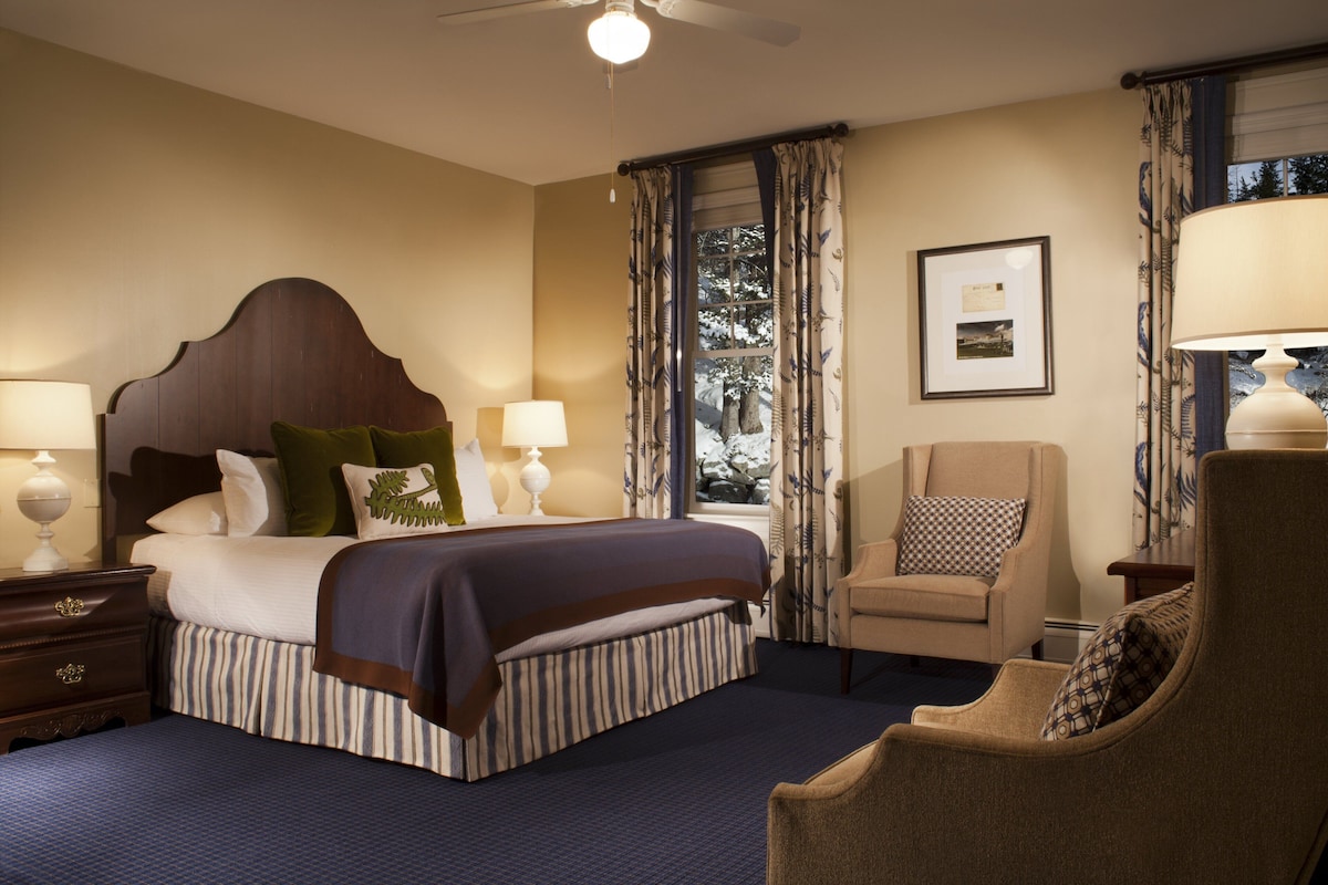 A hotel room with a large bed, a chair, a desk, and a window with curtains.