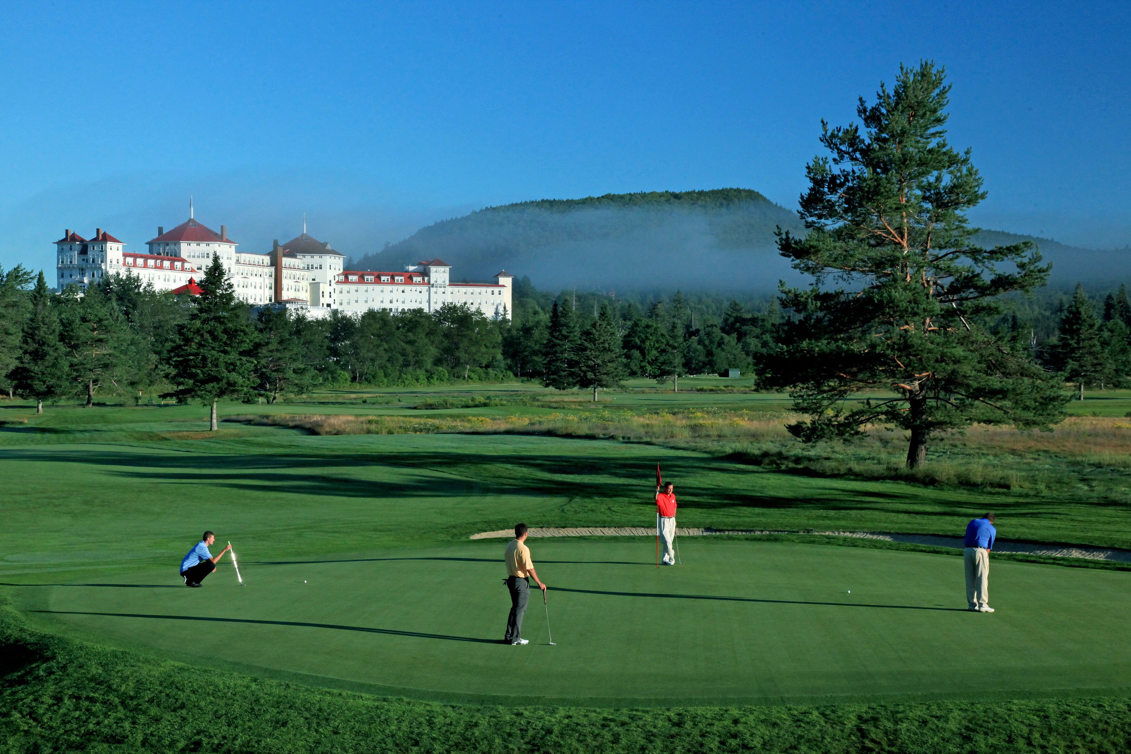 Un campo de golf con jugadores, un edificio grande al fondo y una colina boscosa.