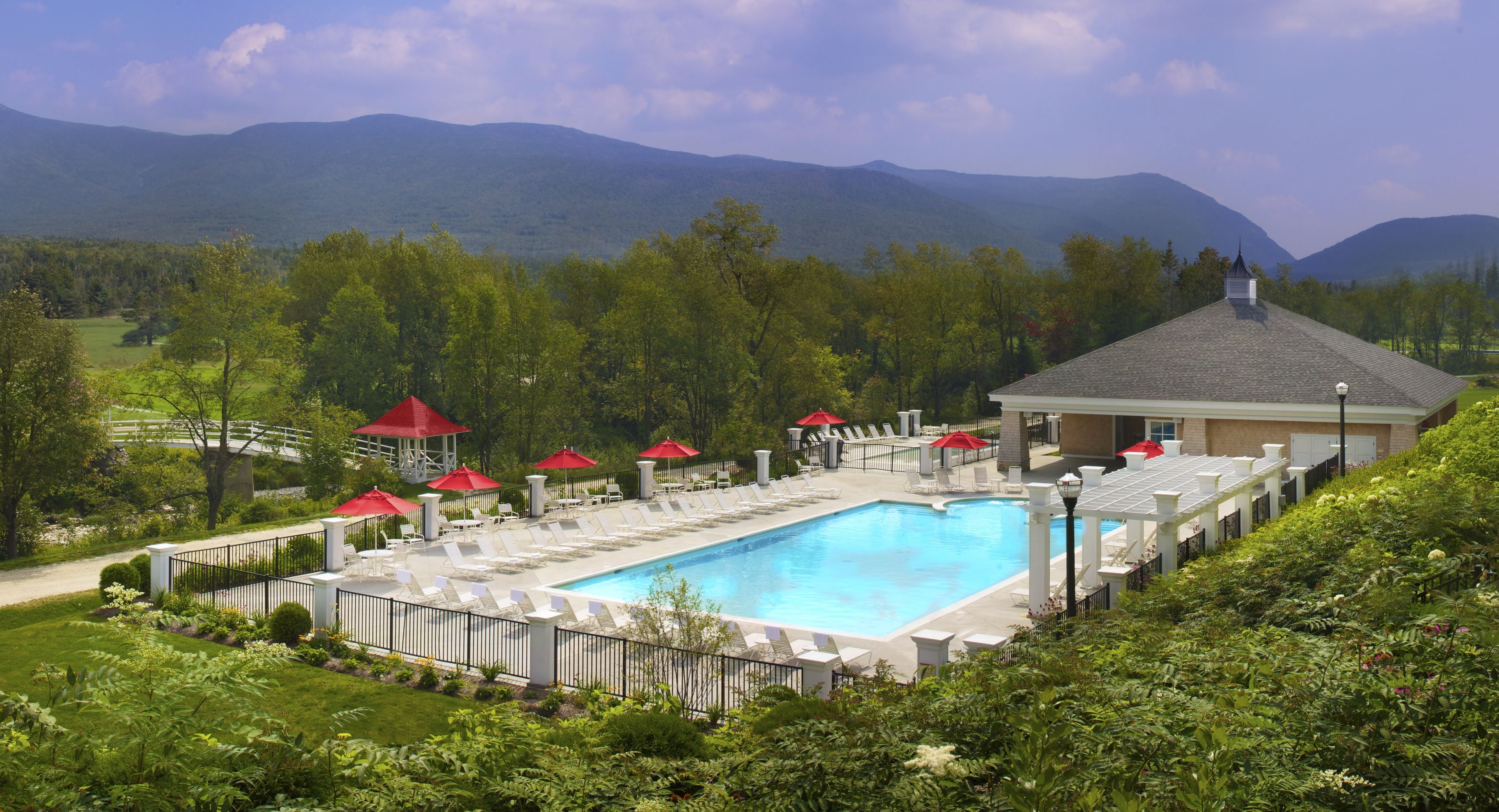 A pool area with lounge chairs and umbrellas, surrounded by greenery and mountains in the background.