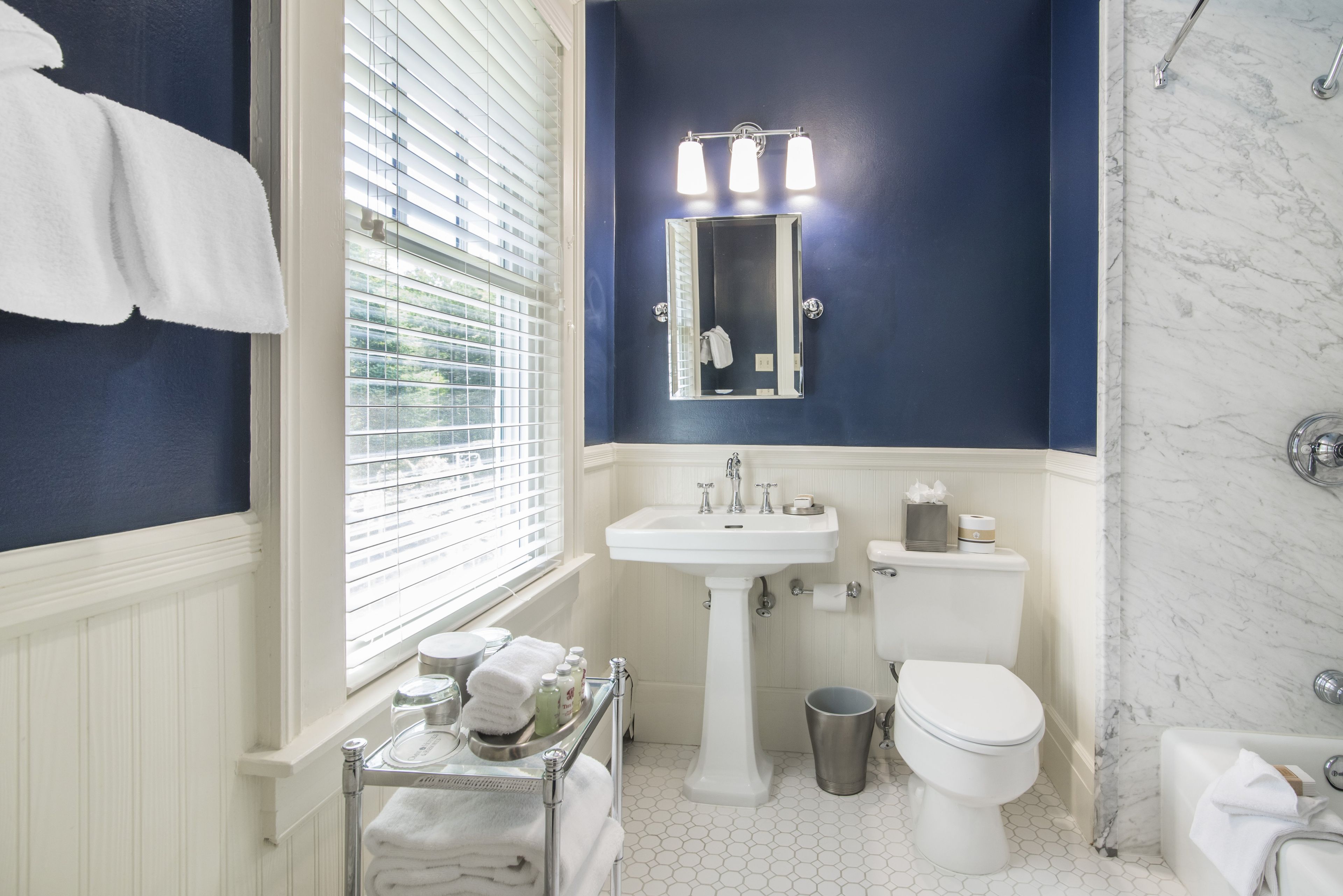 A bathroom with a white sink, a toilet, and a bathtub. There is a window with blinds, a towel rack, and a trash can.