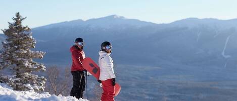 Two individuals on a snowy mountain with a snowboard, a clear sky, and distant mountains.