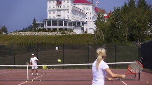A tennis court with a net, a player in a white shirt, and a building with red roofs in the background. - Omni Bretton Arms Inn at Mount Washington (Bretton Woods)