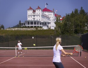 Un court de tennis avec un filet, un joueur portant un maillot blanc, et un bâtiment aux toits rouges en arrière-plan.