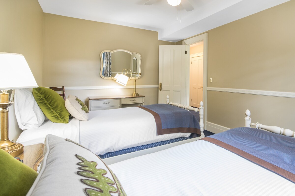 A hotel room with two beds, a bedside table with a lamp, a dresser, and a mirror on the wall.