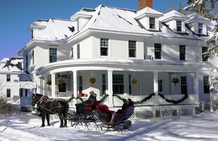 A snow-covered house with a porch, a horse-drawn sleigh, and two people in the sleigh.