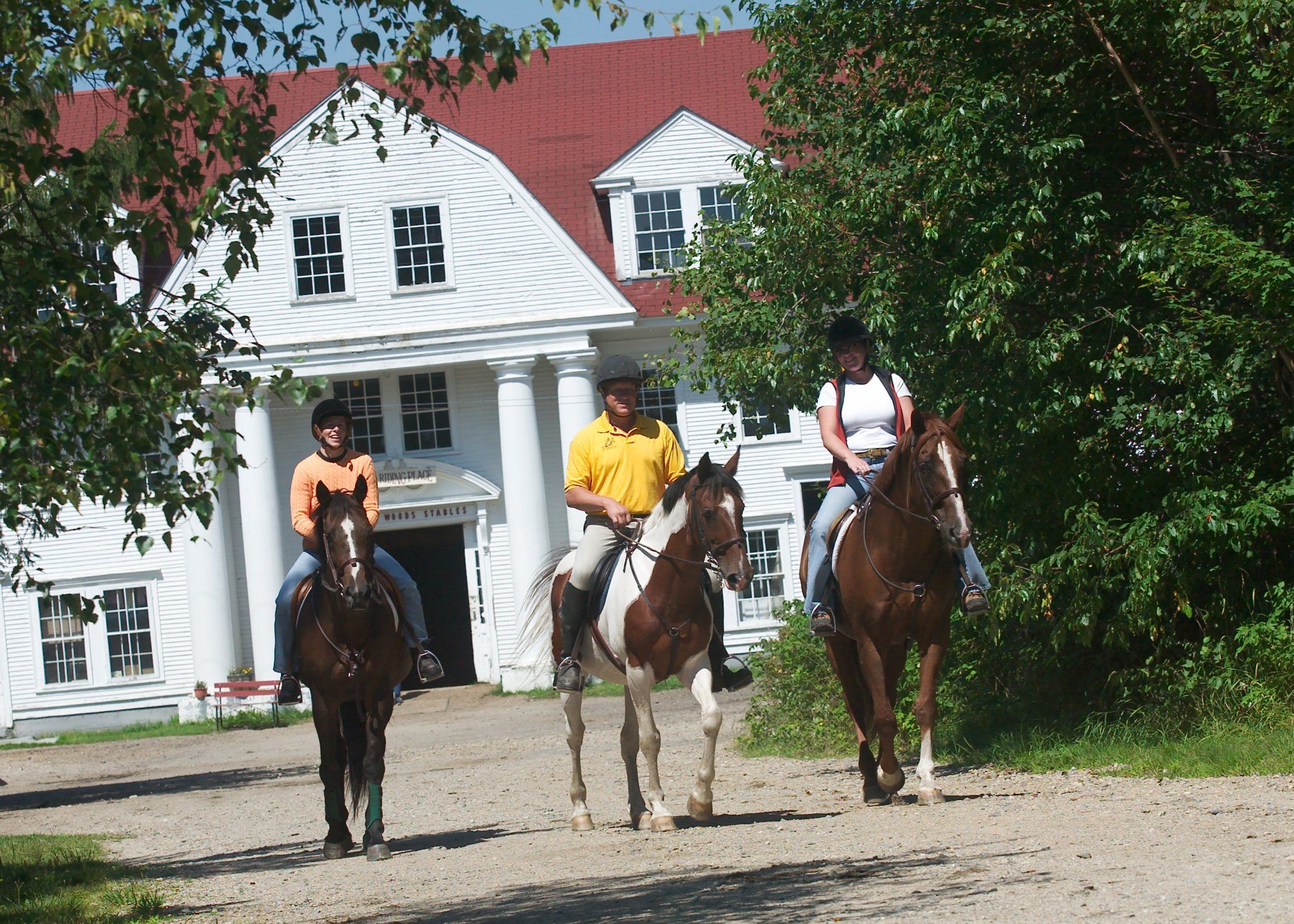 Three people riding horses on a dirt path in front of a white building with a red roof.