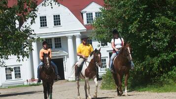 Three people riding horses on a dirt path in front of a white building with a red roof.