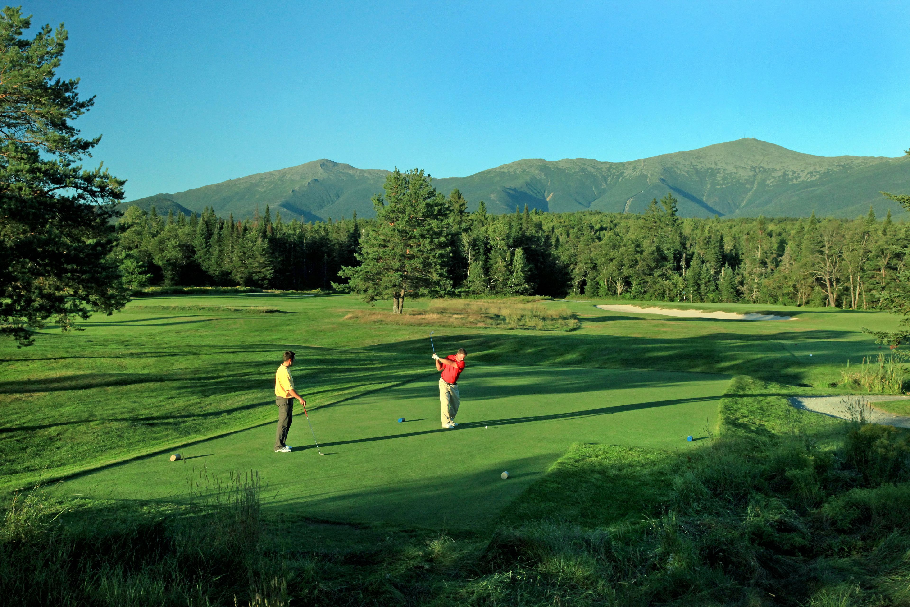 Un campo de golf con cielo despejado, montañas al fondo y un césped bien cuidado.