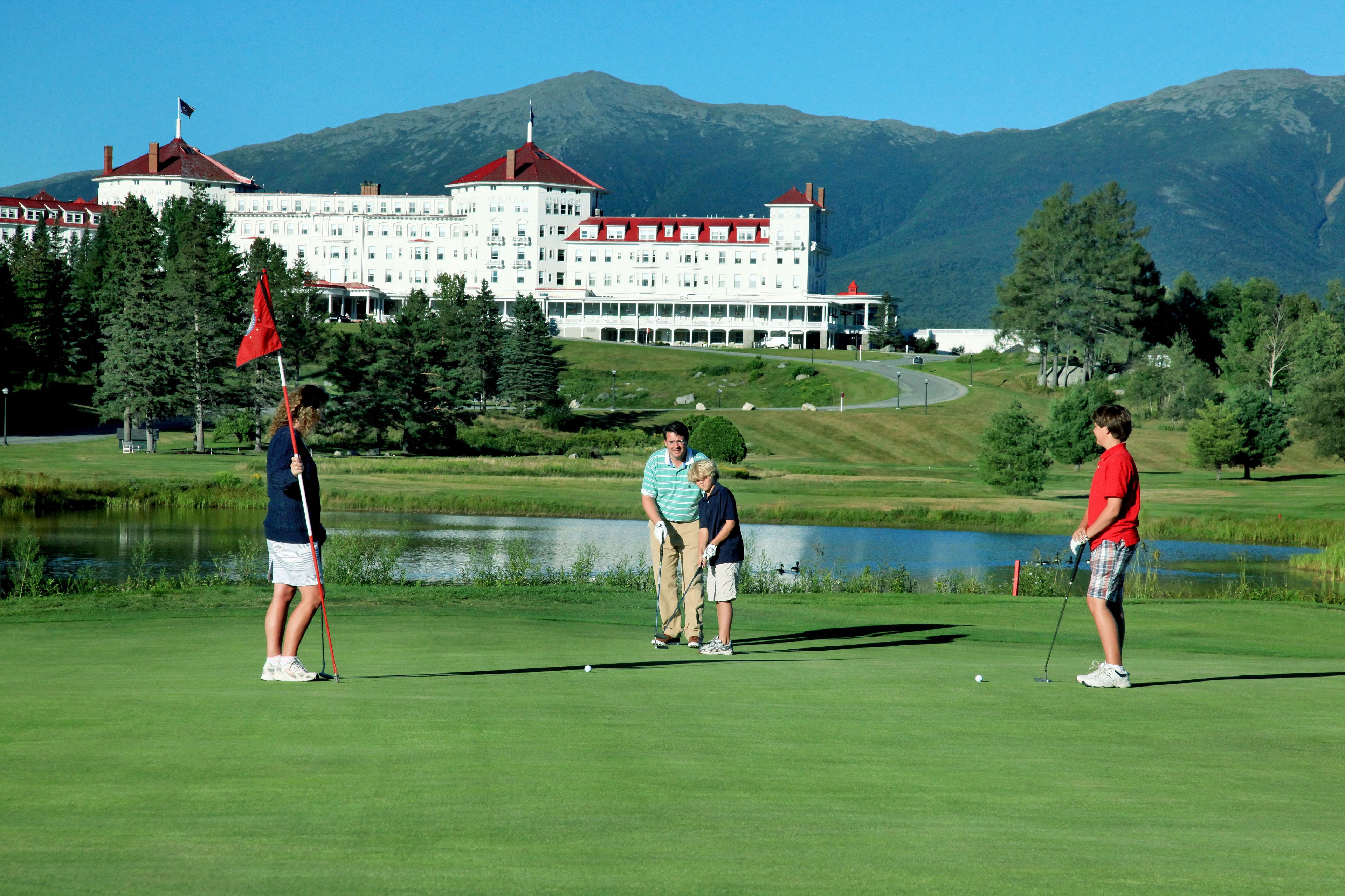 A golf course with a large building in the background, a pond, and a mountain range.