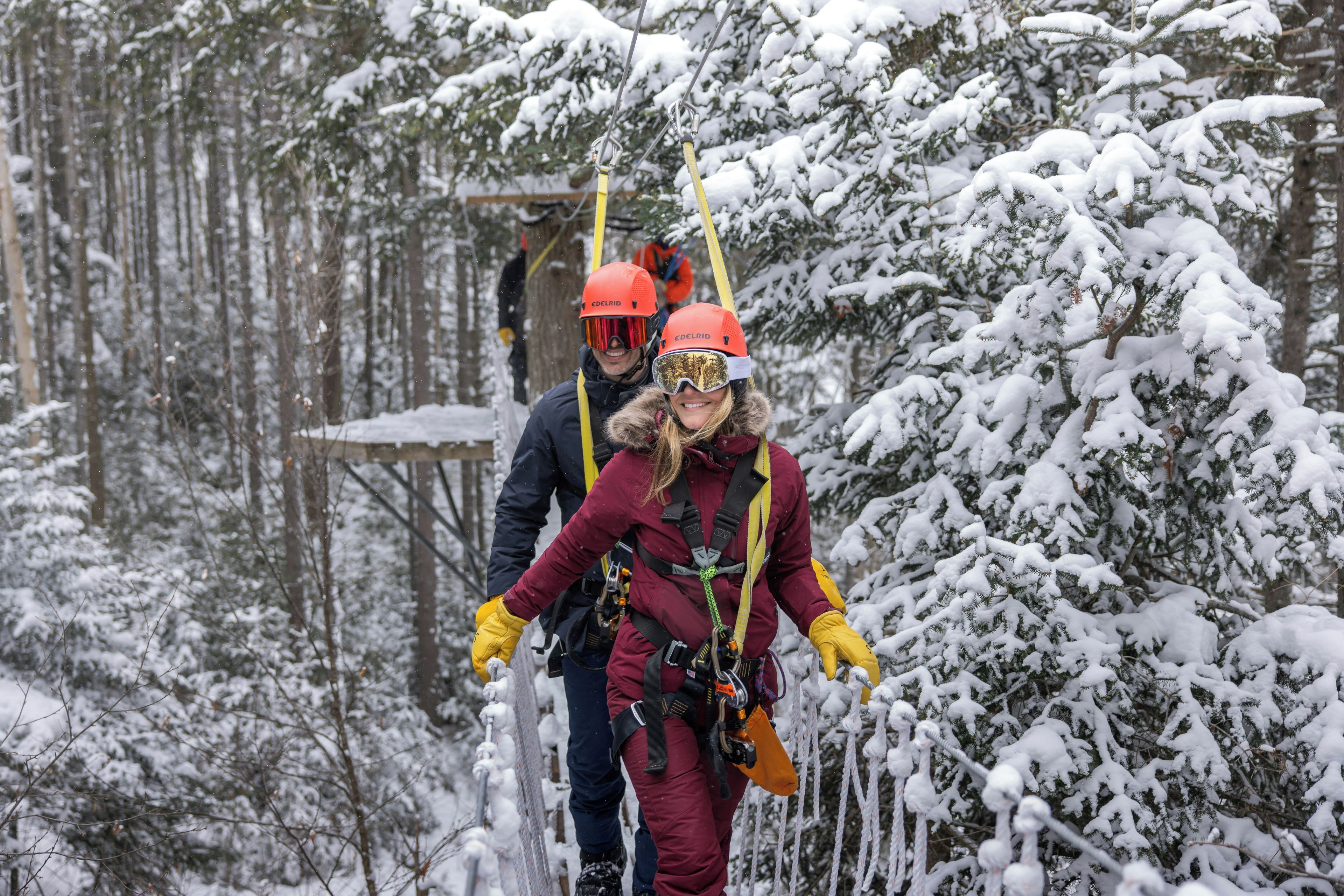 Two individuals in winter gear with safety harnesses, walking through a snowy forest.