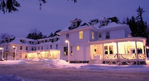 A large, multi-story house with a snow-covered roof and a lit porch.