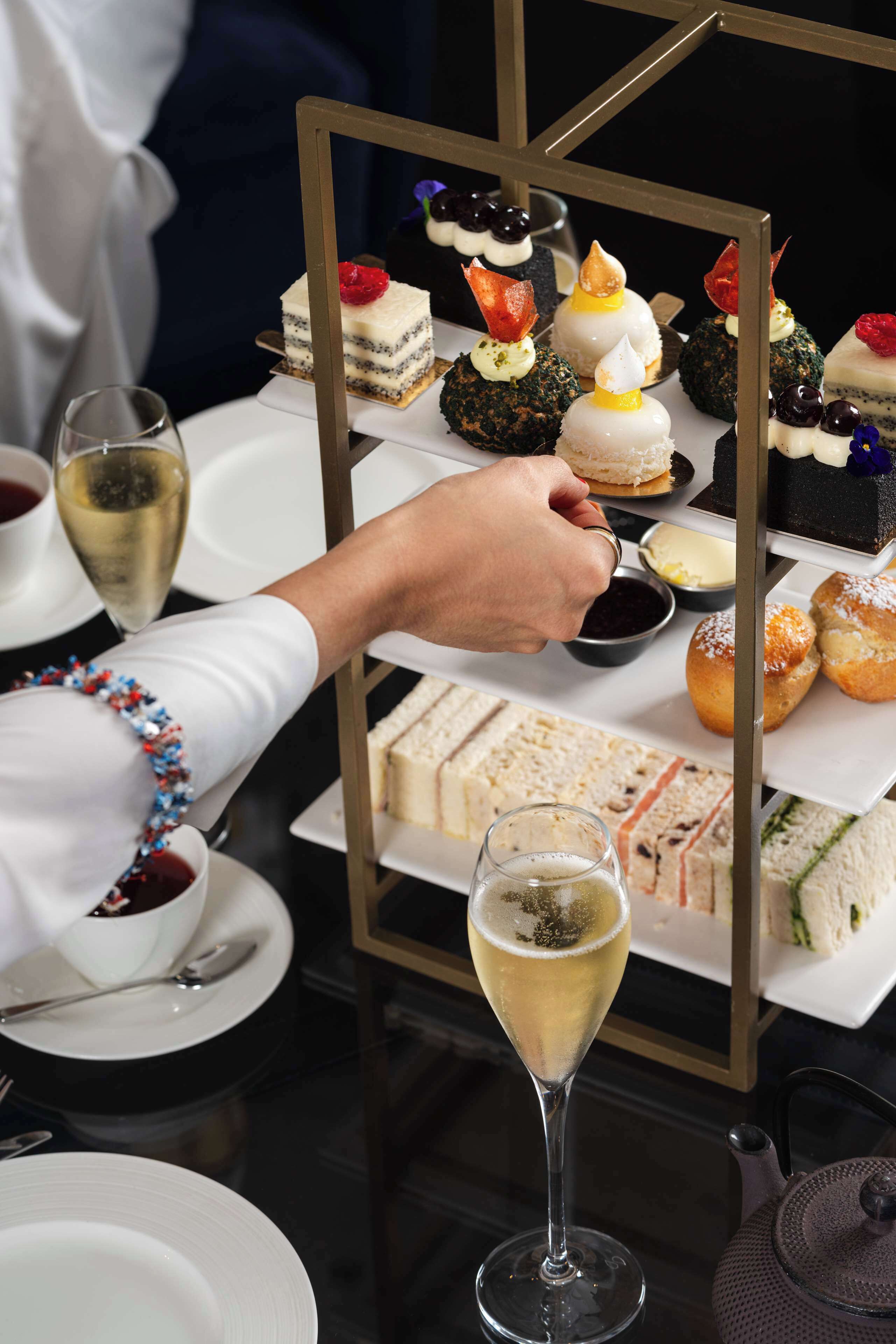 A tray of assorted sandwiches and desserts, a glass of wine, and a teapot on a table.