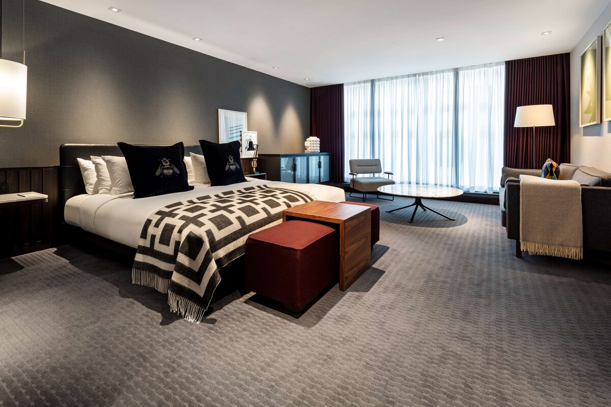 A modern hotel room with a large bed, a desk, a chair, and a view of the city through large windows.
