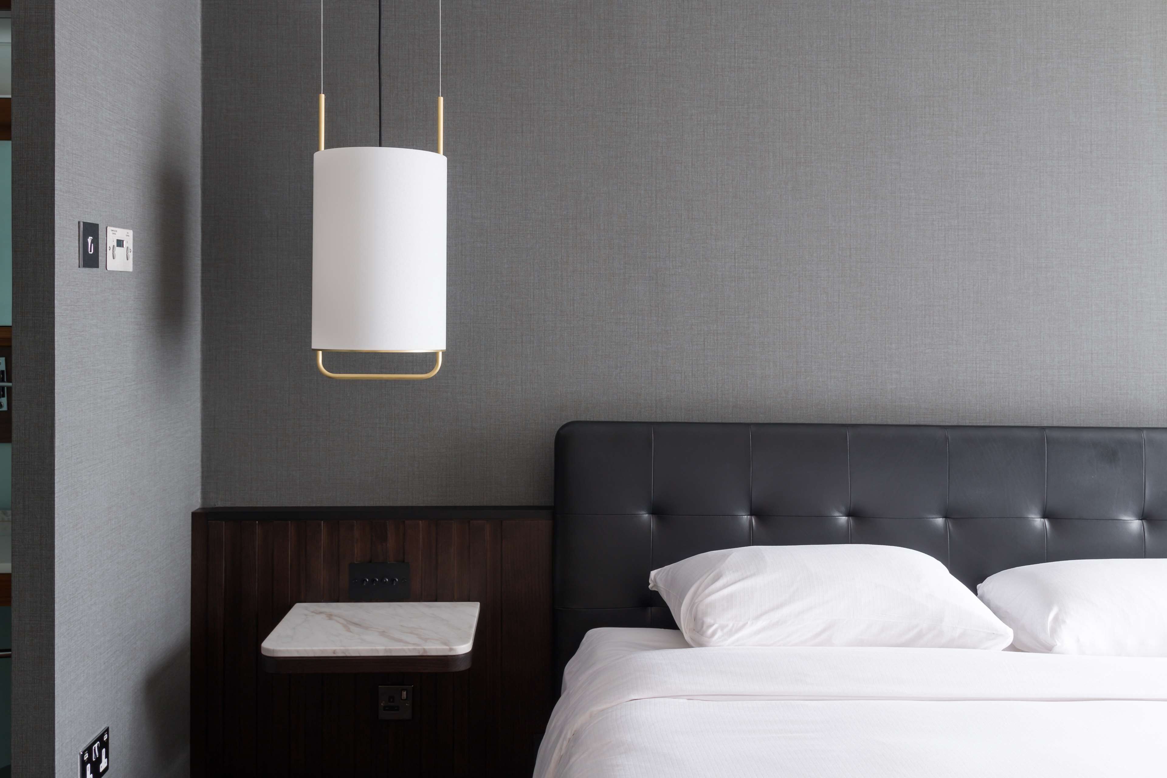 A modern hotel room with a dark headboard, a bedside table with a marble top, and a hanging light fixture.