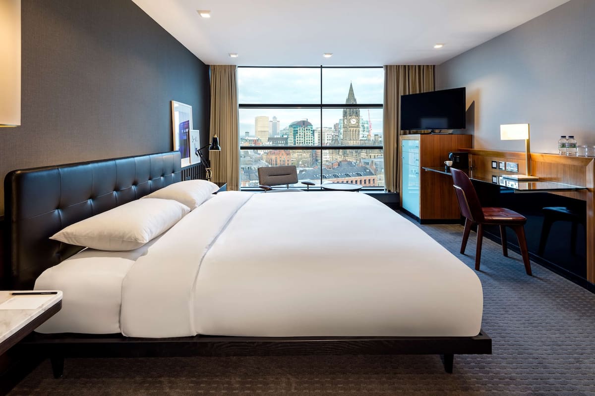 A hotel room with a large bed, a desk, a chair, a TV, and a view of a cityscape.