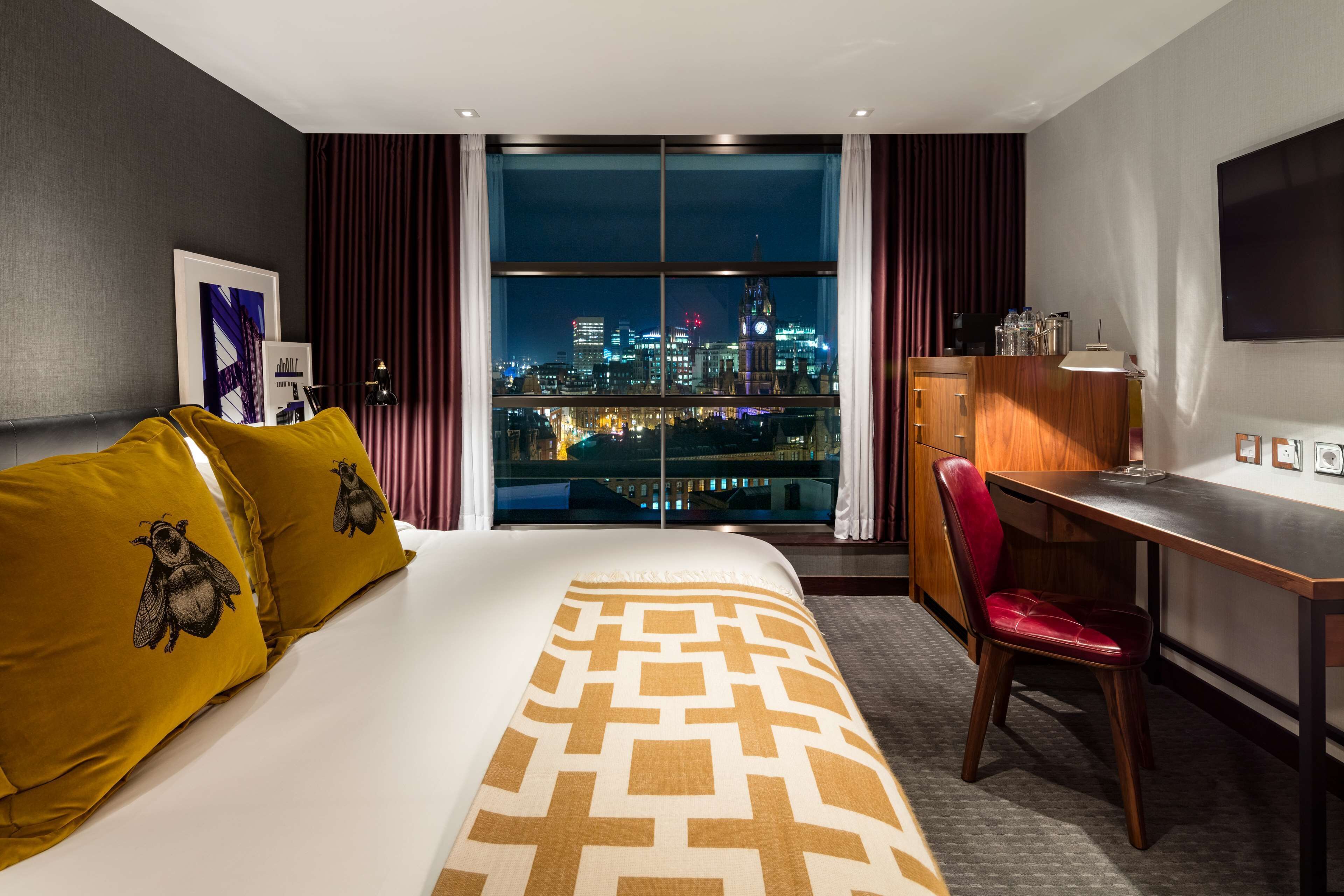 A hotel room with a large bed, a desk, a chair, and a view of a cityscape at night.