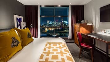 A hotel room with a large bed, a desk, a chair, and a view of a cityscape at night.