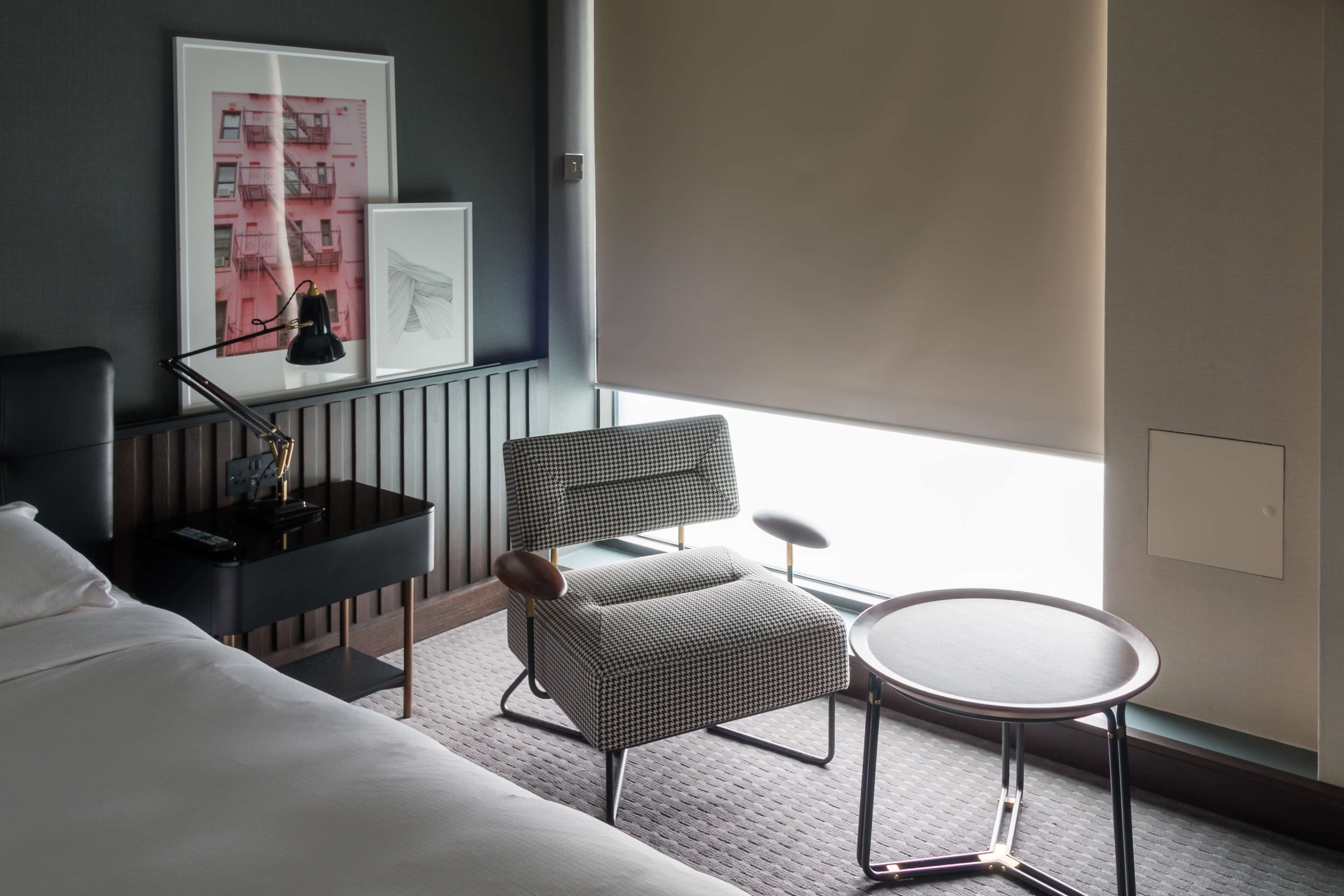A hotel room with a bed, a chair, a small table, and a window with blinds.
